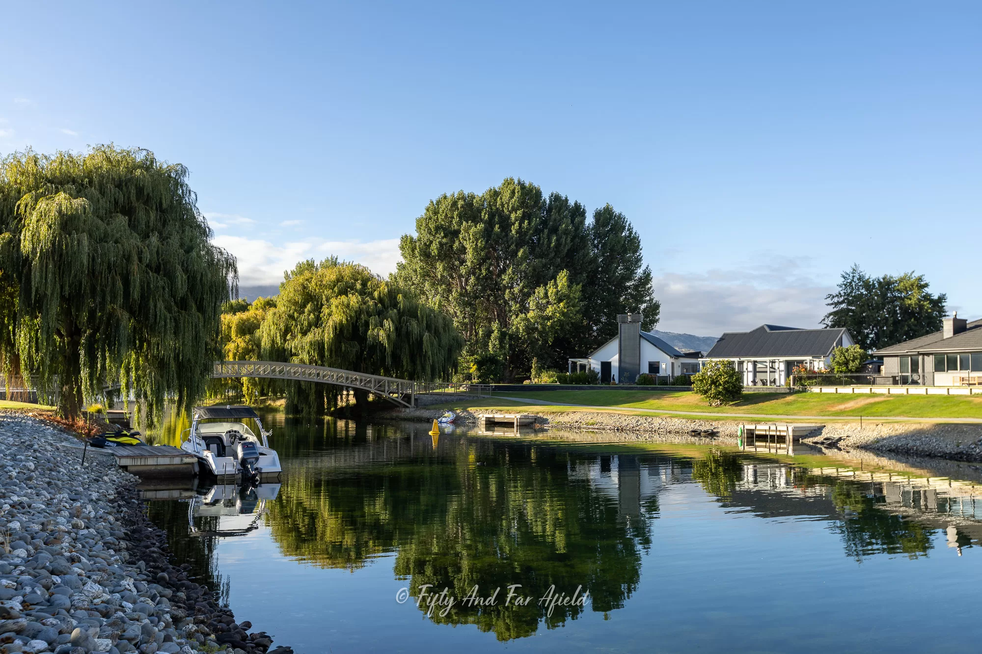 Lakeside canal in Cromwell, New Zealand with moored boats, willow trees, and modern houses reflected in the still water, under a clear blue sky.