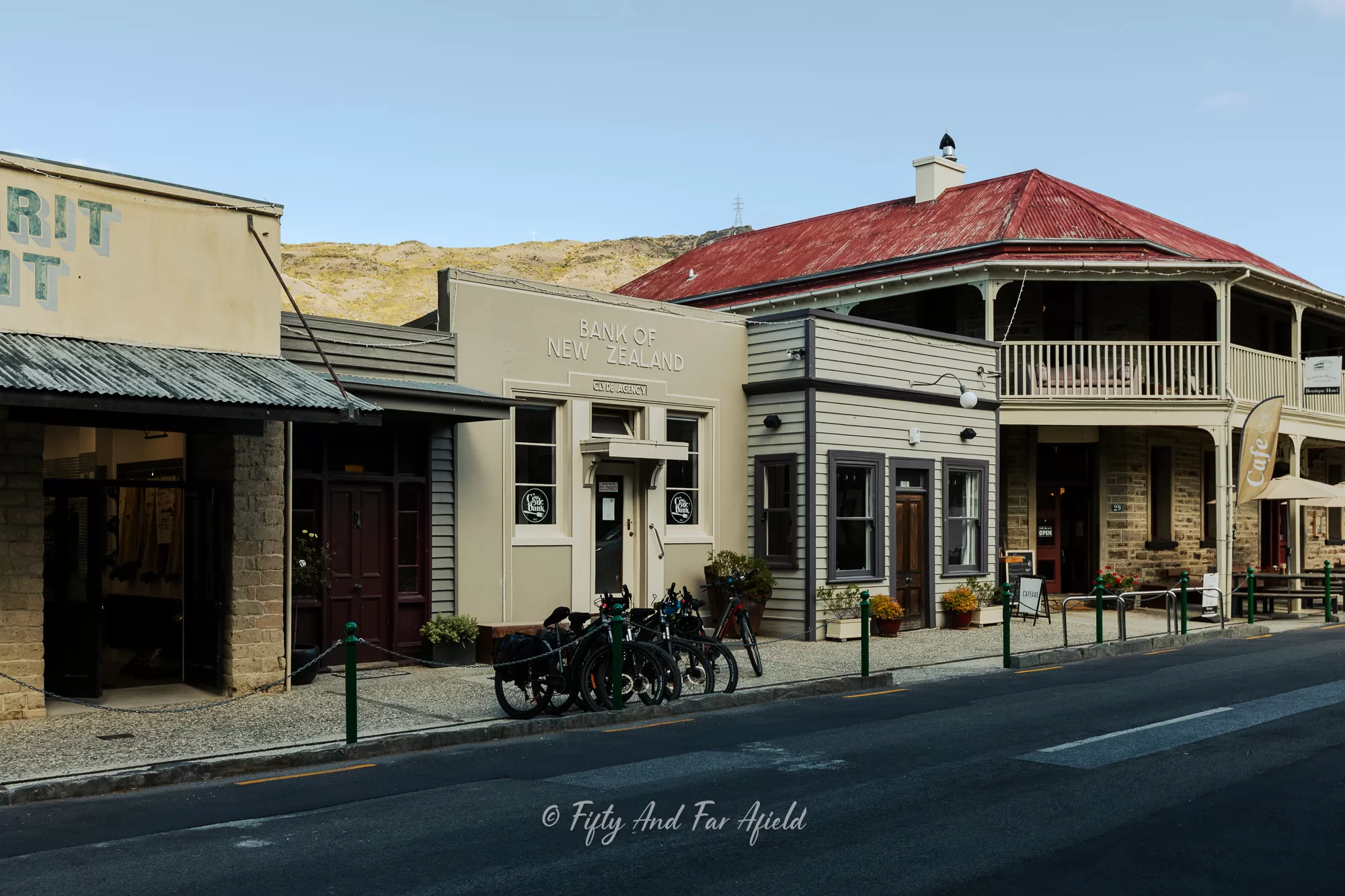 A view of several historic buildings, including the "BANK OF NEW ZEALAND," along Sunderland Street in Clyde, with bicycles parked on the curb and dry hills in the background under a clear sky.