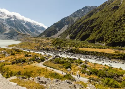 Landscape view of the Hooker Valley Track with the first swing bridge and silhouettes of hikers in the distance on a sunny day in New Zealand’s Southern Alps.