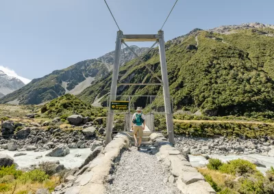 Male hiker with a backpack, viewed from behind, stepping onto the first swing bridge on the Hooker Valley Track in New Zealand’s Southern Alps.