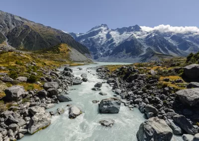 A wide shot of a milky turquoise glacial river winding through a rocky valley in Aoraki/Mount Cook National Park, with snow-capped mountains and scattered clouds in the background.