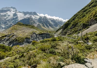 A wide, slightly elevated view of a suspension bridge stretching across a green valley on the Hooker Valley Track in Aoraki/Mount Cook National Park, with snow-covered mountains in the distance and a hiker on a path to the right.