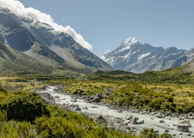 A wide shot of a milky turquoise glacial river flowing through a rocky valley in Aoraki/Mount Cook National Park, with snow-capped mountains and scattered clouds in the background.