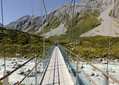 A perspective view from the entrance of a long suspension bridge with a wooden walkway and metal railings, stretching over a milky turquoise river in a rocky valley with steep mountains on either side.
