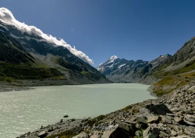 A wide view of Hooker Lake, a milky turquoise glacial lake, surrounded by rocky terrain and towering mountains, with the snow-capped Mount Cook visible in the center against a blue sky with some clouds.