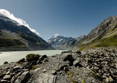 A wide view of Hooker Lake, a milky turquoise glacial lake, surrounded by rocky terrain and towering mountains, with the snow-capped Mount Cook visible in the center against a blue sky with some clouds.