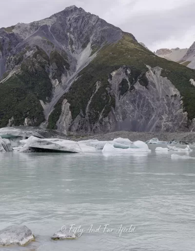 A view of Tasman Lake with several white and grey icebergs floating on its milky turquoise surface, set against a backdrop of tall, rugged mountains under a cloudy sky in Aoraki/Mount Cook National Park.