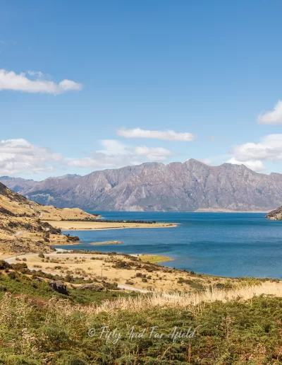 A wide panoramic view of the deep blue waters of Lake Hāwea surrounded by dry, golden-brown hills and mountains under a bright blue sky with scattered clouds, as seen from a lookout point.