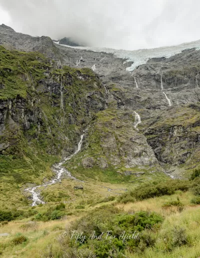 A view of a glacier situated high on rugged mountains with multiple waterfalls flowing down, as seen from the Rob Roy's Glacier Track and lookout in Mt Aspiring National Park, under an overcast sky.