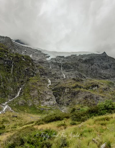 A view of a glacier situated high on rugged mountains with multiple waterfalls flowing down, as seen from the Rob Roy's Glacier Track and lookout in Mt Aspiring National Park, under an overcast sky.