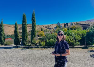 Anna, a travel blogger in sunglasses and a striped shirt, stands on a gravel path at a vineyard with rows of grapevines, cypress trees, and rolling hills under a clear blue sky.