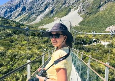 Anna, a travel blogger wearing a yellow shirt, hat, and sunglasses with a camera, stands on a suspension bridge with a wooden walkway and wire sides, overlooking a green valley with mountains in the background under a blue sky.
