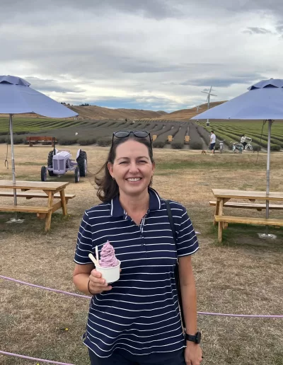 Anna, a travel blogger in a striped shirt with sunglasses on her head, holds a cup of lavender ice cream at the NZ Alpine Lavender farm, with rows of lavender, picnic tables with purple umbrellas, and vintage farm equipment in the background under a cloudy sky.