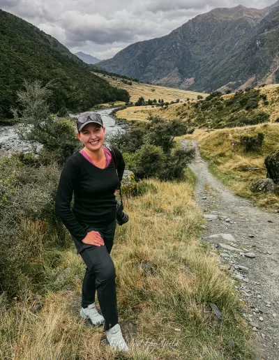 Anna, a travel blogger, stands on a track in a valley with a river to the left and mountains on either side under a cloudy sky, on Rob Roy's Glacier Track in Mt Aspiring National Park.
