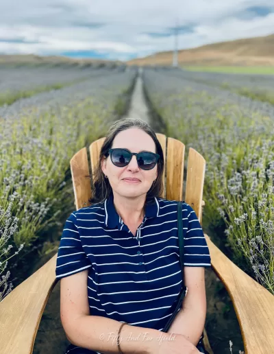 Anna Jackowska, wearing sunglasses and a striped shirt, sits in a large wooden chair surrounded by rows of blooming purple lavender plants at a farm near Lake Tekapo, under an overcast sky.