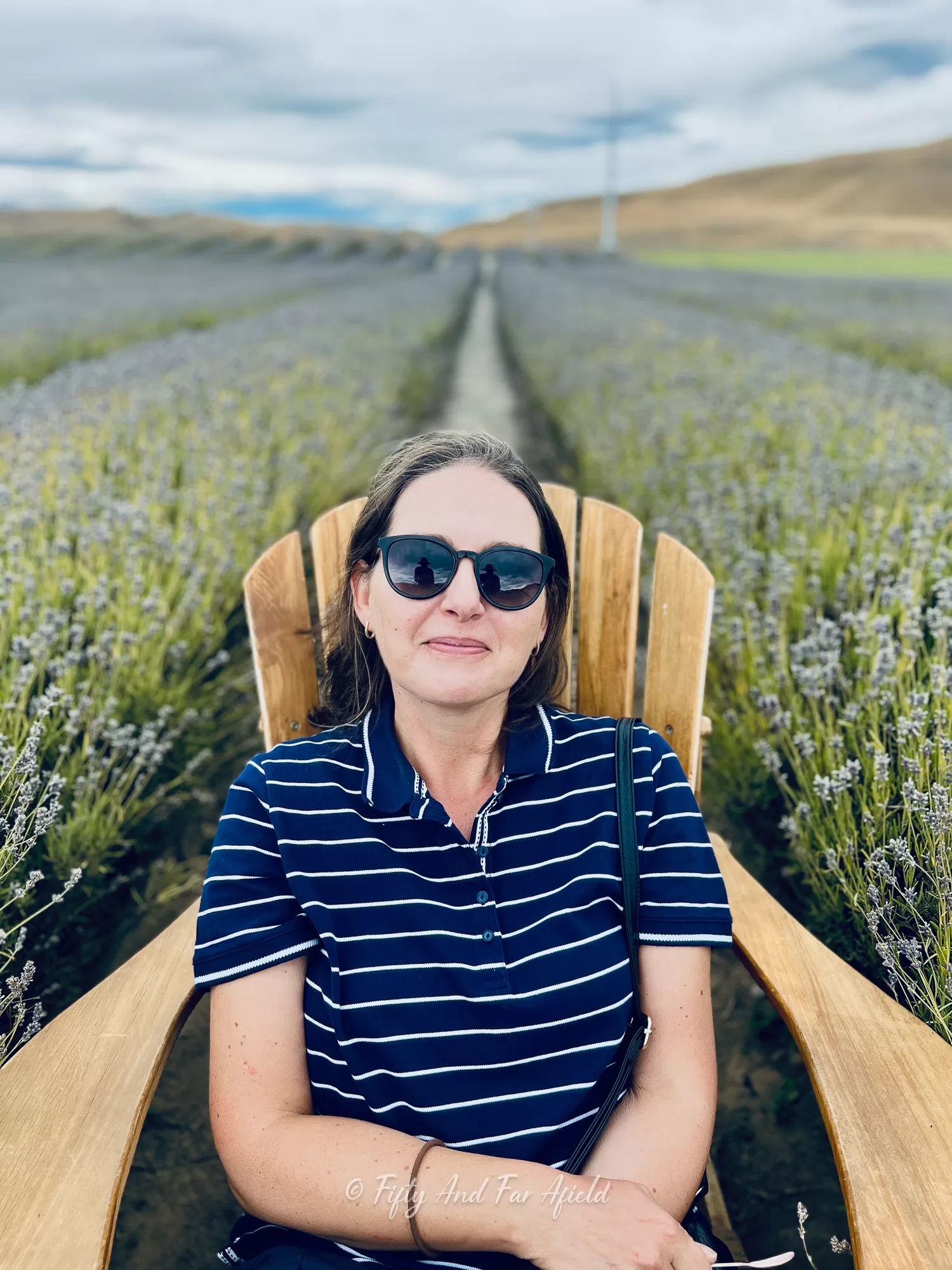 Anna Jackowska, wearing sunglasses and a striped shirt, sits in a large wooden chair surrounded by rows of blooming purple lavender plants at a farm near Lake Tekapo, under an overcast sky.
