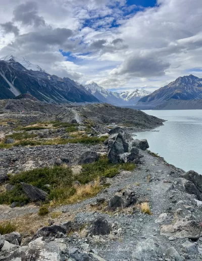 A view of a rocky path along a ridge overlooking Tasman Lake, with snow-capped mountains in the background under a partly cloudy sky.