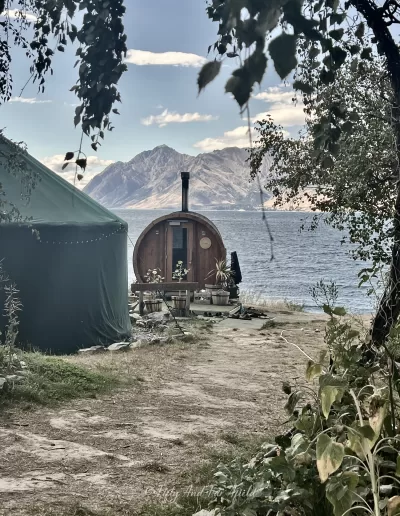 A barrel-shaped wooden sauna and a dark green tent are located near the blue waters of Lake Hāwea, with rugged mountains in the background under a partly cloudy sky.