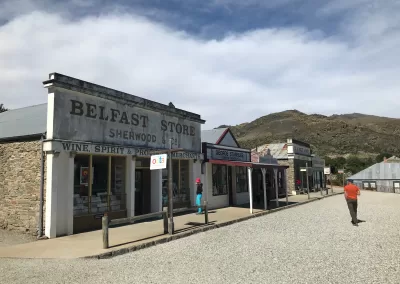A view of the Belfast Store and other historic buildings along a gravel street in the Cromwell Historic Precinct, with two people present and dry hills in the background under a partly cloudy sky.