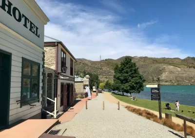 A view of a gravel path alongside the turquoise waters of Lake Dunstan in the Cromwell Historic Precinct, with historic buildings on the left and a grassy area with people and trees on the right, under a sunny sky.