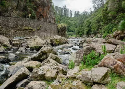 A wide shot of a river with many large boulders and rocks in its bed, flowing through a gorge with steep, green, forested hillsides and a stone wall on the left supporting a road.