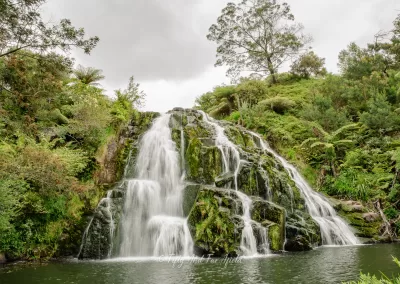 A wide shot of a multi-tiered waterfall cascading over dark, mossy rocks into a calm pool of water at the bottom. The waterfall is surrounded by dense green native vegetation and trees under a cloudy sky.