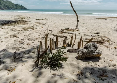 A small, whimsical structure made of upright sticks and driftwood stands on a sandy beach, with a rock and some greenery nearby. In the background, the wide sandy beach meets the ocean with gentle waves under a clear sky.