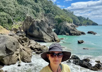 A woman with dark sunglasses and a wide-brimmed grey hat, wearing a yellow t-shirt, smiles directly at the camera. Behind her is a picturesque rocky beach with turquoise ocean water and a steep, green, tree-covered hillside under a bright blue sky with white clouds.