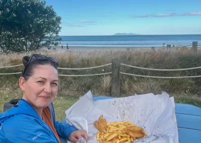 A woman with dark hair pulled back and sunglasses on her head, wearing a blue jacket over an orange top, smiles while sitting at a blue picnic table. In front of her is a large serving of fish and chips wrapped in paper. Behind her is a sandy beach, the ocean, and distant land under a blue sky with seagulls flying.