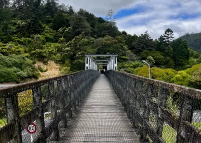 A long, narrow suspension bridge made of wood and metal mesh, extending straight into the distance towards a forested hillside. A red and white circular speed limit sign with "40" on it is attached to the left railing. The sky is cloudy with patches of blue.