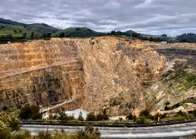 A very large open-pit mine with many visible terraced levels, showing shades of brown, orange, and grey rock. The mine stretches across the middle of the image under a cloudy sky, with green hills visible in the background and some low scrub in the foreground.