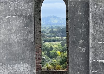 A view through a tall, arched window opening in a cracked, grey concrete wall. The window frames a landscape of rolling green hills, some trees, and distant houses under a cloudy sky.