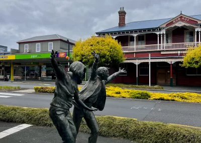 A bronze sculpture of two children with their arms raised in a happy or excited gesture, standing on a light-colored plinth in the foreground. Behind them is a street, and across the street are a red historic building with "ROB ROY" on it and a green and white modern building. Yellow-leafed trees are visible.