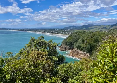 A wide panoramic view of a long, curving white sand beach with turquoise ocean water, seen from a high vantage point. The foreground is filled with dark green native bush. Distant green hills are visible on the horizon under a bright blue sky with scattered white clouds.