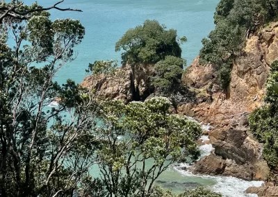 A high-angle view overlooking a secluded bay with brilliant turquoise water. Rocky cliffs and outcrops covered in green trees frame the bay, with waves breaking on the shore. In the foreground, more green trees with white blossoms are visible, and distant islands can be seen on the horizon under a blue sky with scattered clouds.